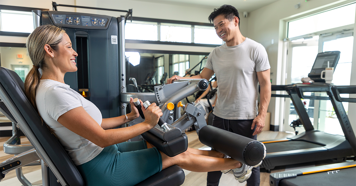 Woman performs leg extension exercise on gym machine while a trainer stands nearby smiling.