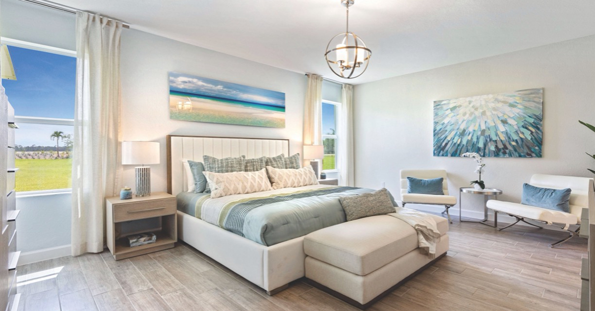 Bright staged bedroom with upholstered bed, lamps, artwork, and large windows overlooking grass