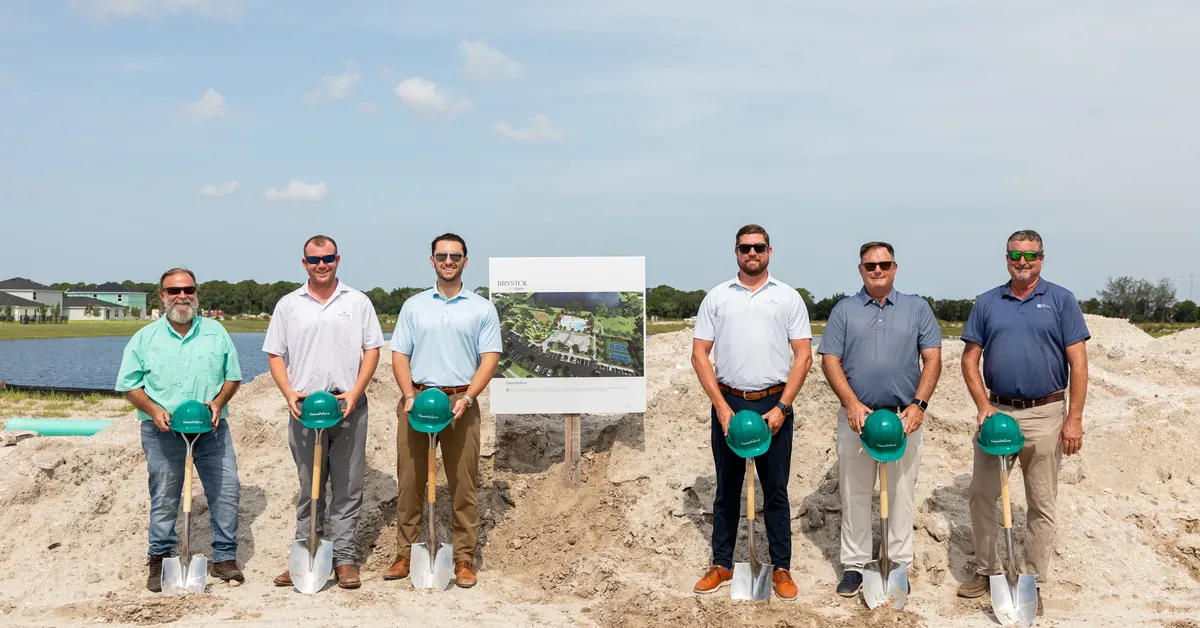 Six executives hold shovels and green hard hats at a real estate groundbreaking ceremony beside an architectural rendering board.