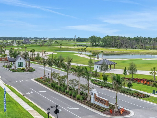 Aerial view of a golf course community entrance with a stone monument sign, palm-lined roadway, and golf course with ponds in the background.