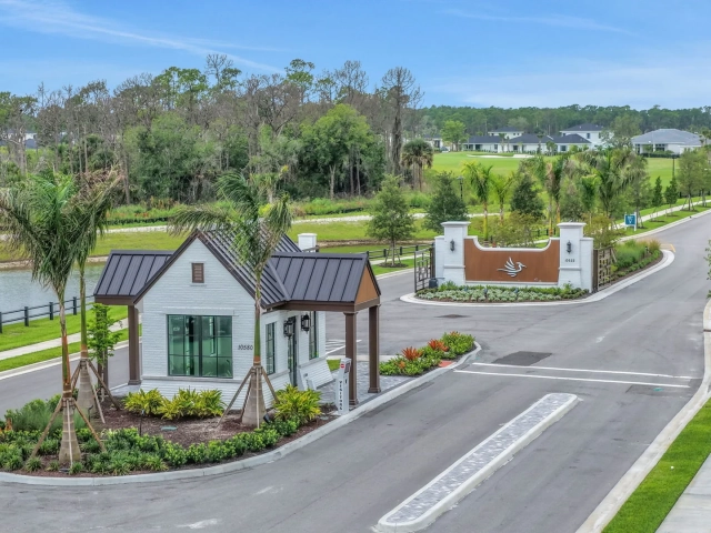Aerial view of a white guard house and a curved entry gate with a bird logo, next to a lake and a road lined with palm trees.