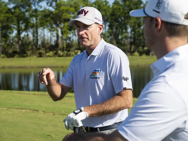 A man in an Ally golf cap and striped polo shirt gesturing while talking with another golfer in a white cap on a golf course.