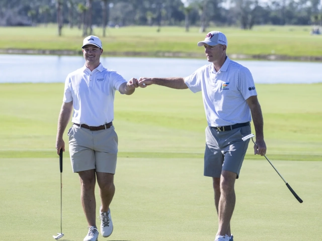 Two men in golf caps and polo shirts fist-bumping while walking across a putting green, with a lake in the background.