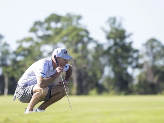 A man in a white golf cap and striped polo shirt crouching down to line up a putt on a golf green.