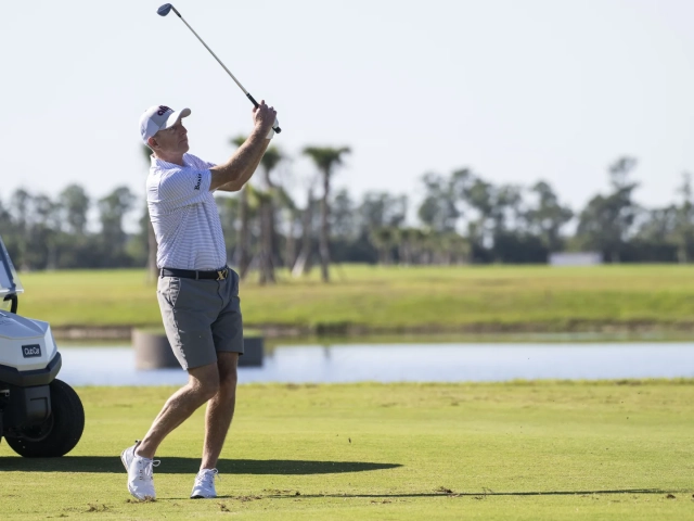 A man in a white golf cap and striped polo shirt following through on a golf swing beside a white golf cart on a course.