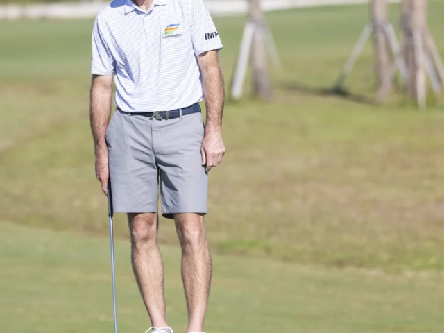 A man in a white golf cap and striped polo shirt standing on a putting green watching a golf ball roll toward the hole.