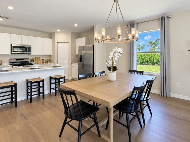 Dining table with six black chairs beneath a chandelier, next to a white kitchen with stainless steel appliances.