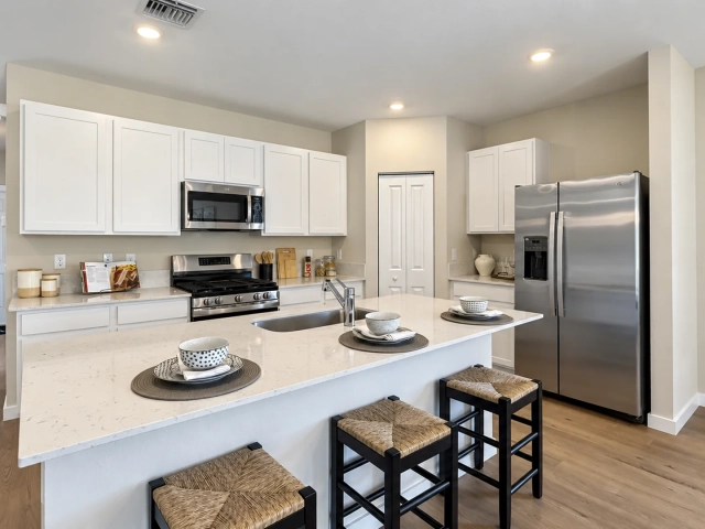 White kitchen with a long quartz island, three woven bar stools, and stainless steel refrigerator and range.