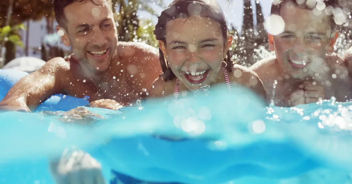 Family laughing and splashing together in a bright blue swimming pool on a sunny day, surrounded by tropical palm trees.