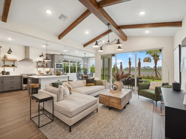 Open living room with exposed wood beam ceiling, a beige sectional sofa, and an open kitchen with gray cabinets in the background.
