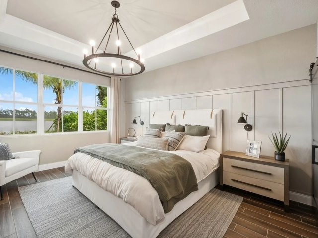 Primary bedroom with a tray ceiling, a wagon-wheel chandelier, an upholstered bed with green bedding, and a large window overlooking a lake.