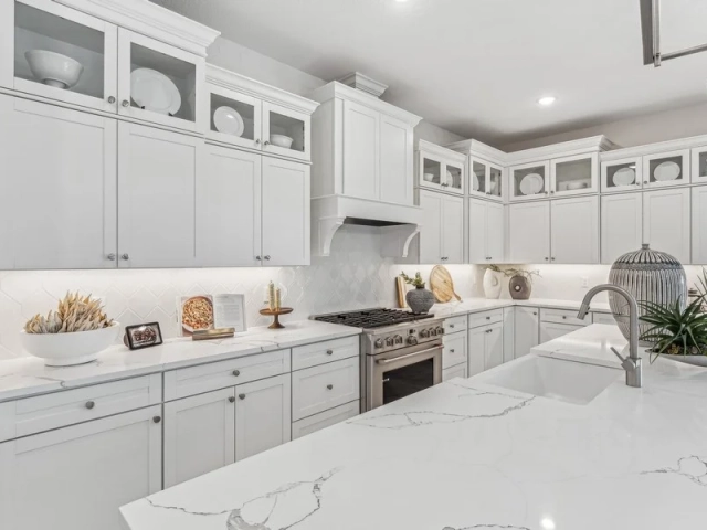 Kitchen with light gray cabinets, glass-front upper cabinets displaying dishware, a stainless steel range, and a white quartz island.