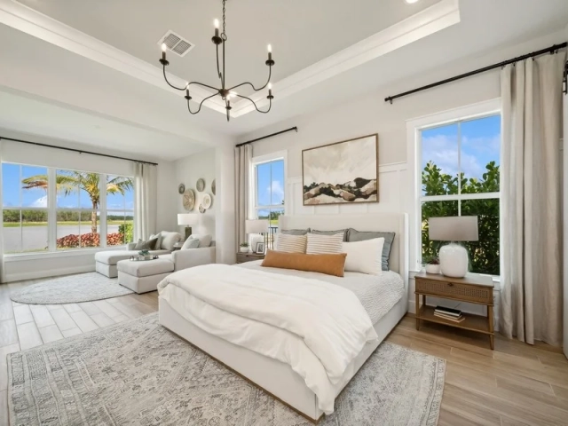 Primary bedroom with a white upholstered bed, a wrought iron chandelier, and a sitting area with a chaise lounge near a window overlooking water.