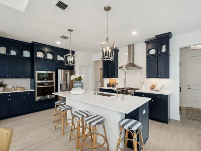 Navy blue kitchen with a large white island, five striped rattan barstools, and stainless steel wall oven and refrigerator.