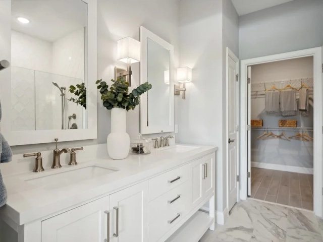 Bathroom with a white dual vanity, framed mirrors, sconce lighting, and a doorway leading into a walk-in closet.