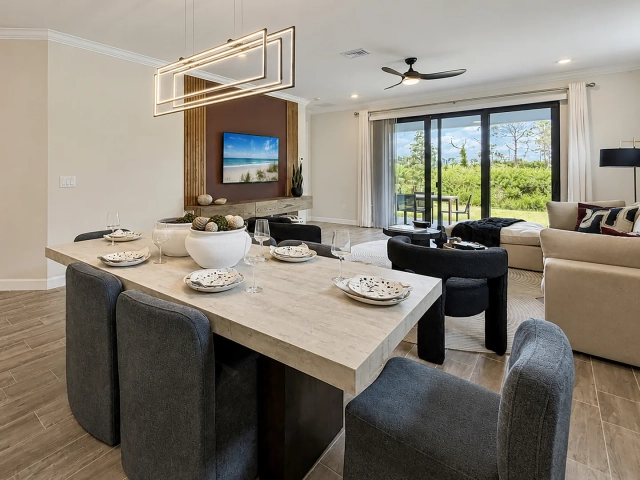 Dining table with dark upholstered chairs and place settings beneath a geometric chandelier, with a wall-mounted TV nearby.