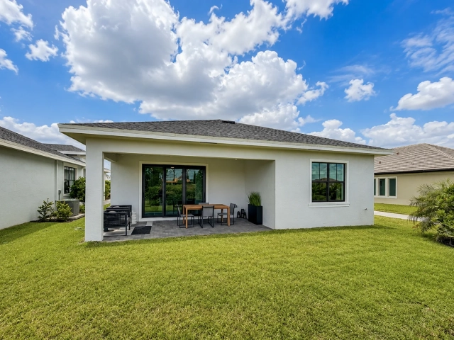 Covered patio with an outdoor dining table and dark lounge chairs, opening onto a spacious backyard lawn.