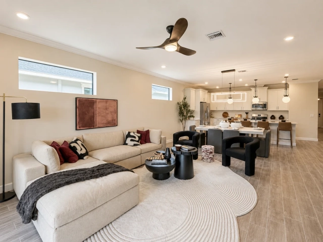 Living room with a cream sectional sofa, abstract wall art, and an open kitchen with globe pendant lights in the background.