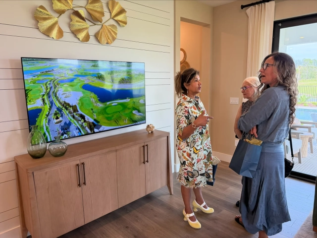 A woman pointing at a wall-mounted TV showing an aerial view of a golf course community while talking with two guests.