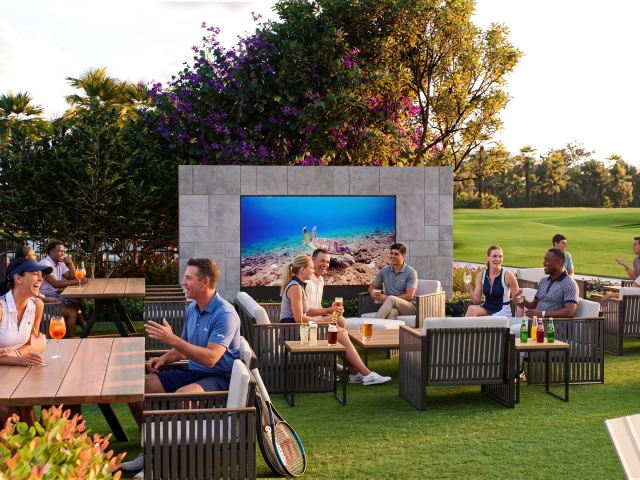 Rendering of people relaxing on outdoor lounge furniture around a large mounted TV screen on a grassy lawn, golf course in the background.