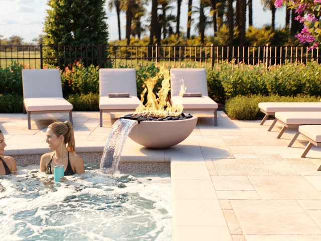 Rendering of two women relaxing in a pool beside a stone fire bowl with a water spout, surrounded by lounge chairs and palm trees.