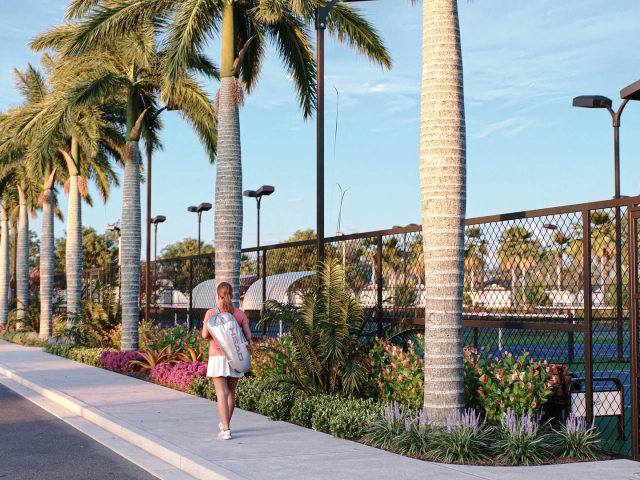Rendering of a woman carrying a tennis bag walking along a palm-lined path beside fenced tennis and pickleball courts.