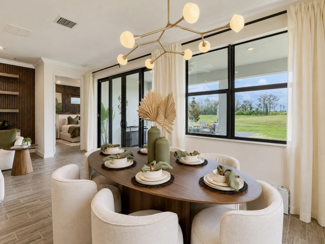 Oval wood dining table set for six with cream chairs, beneath a gold branching chandelier and large windows.