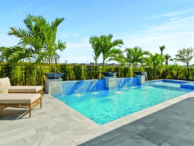 Backyard pool with a raised wall featuring five cascading water spouts, lounge chairs, and palm trees along the fence line.