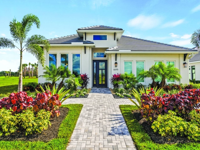 Single-story white model home with a tiled roof, dark front door, and dense red and green landscaping along a paver walkway, address 6695.