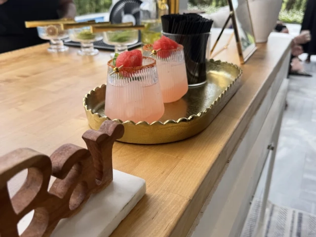 Pink cocktails with watermelon garnish on a gold tray beside a floral arrangement and brass beverage tap at an event bar.