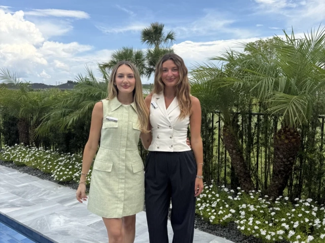 Two women smiling together outdoors beside a pool, one in a sage green dress and one in a white vest with dark trousers.