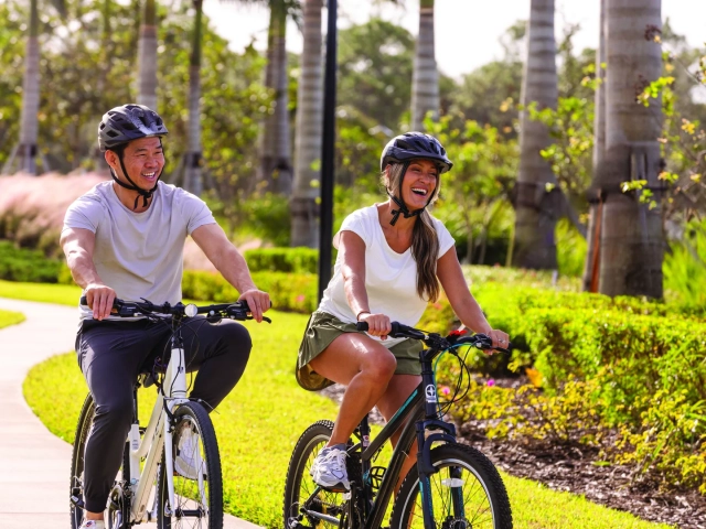 A man and woman wearing bike helmets smiling while riding bicycles along a landscaped path lined with palm trees.