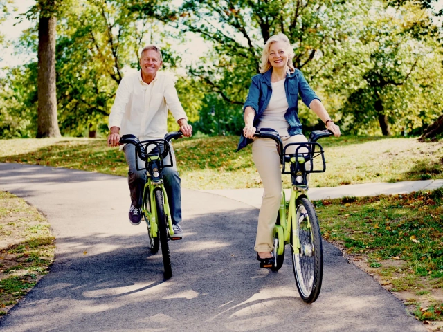 A man and woman smiling while riding bicycles along a paved path lined with trees.