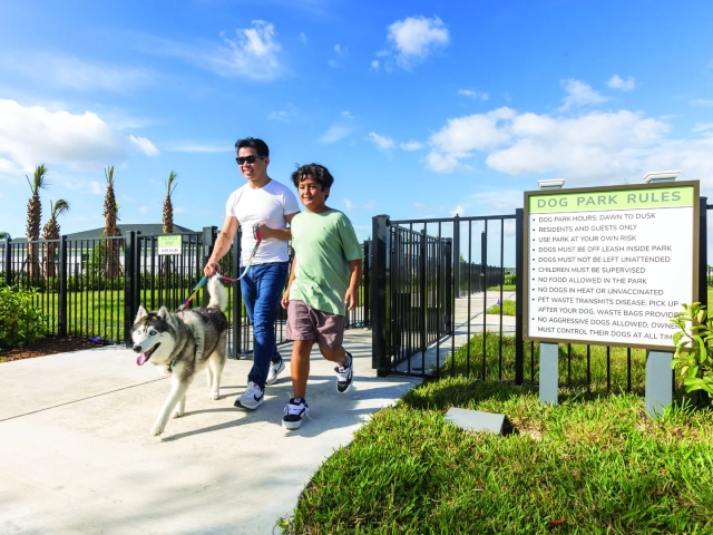 A man and a boy walking a husky on a leash into a fenced dog park, with a dog park rules sign nearby.