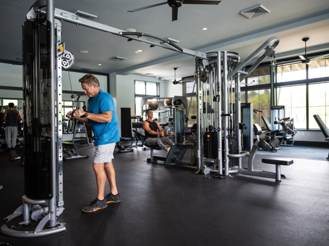 A man using a cable pulley weight machine in a bright fitness center, with another man exercising on a rowing machine in the background.