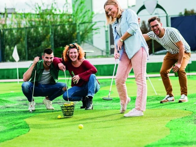 A group of four friends laughing together while playing mini golf, with one woman mid-putt on the green.