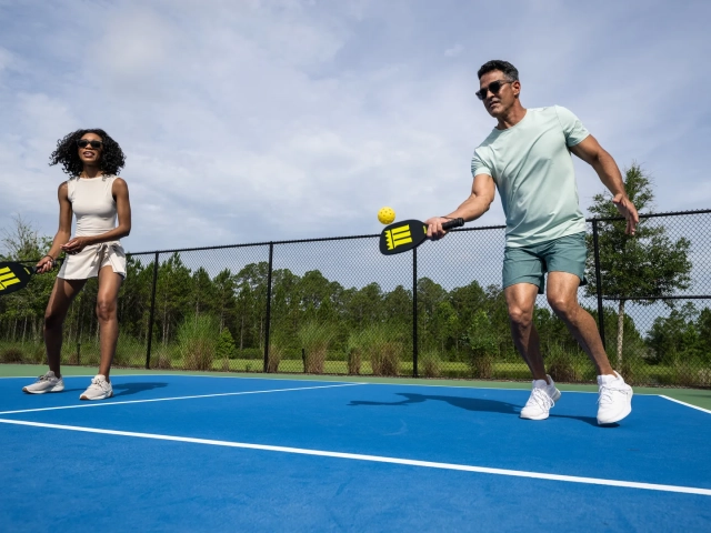 A man and woman playing pickleball on a blue and green court, with the man mid-swing hitting a yellow ball.