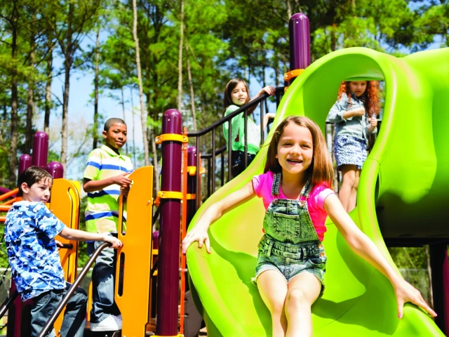 A group of children playing on a colorful playground, with one girl sliding down a green tube slide.