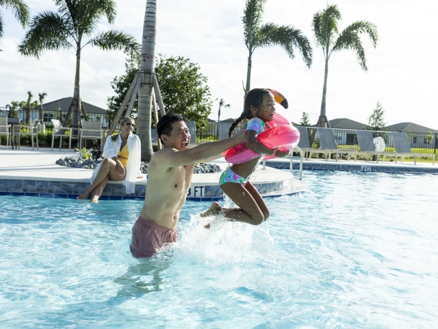 A man lifting a laughing young girl with a flamingo pool float out of a swimming pool, with a woman sitting poolside nearby.
