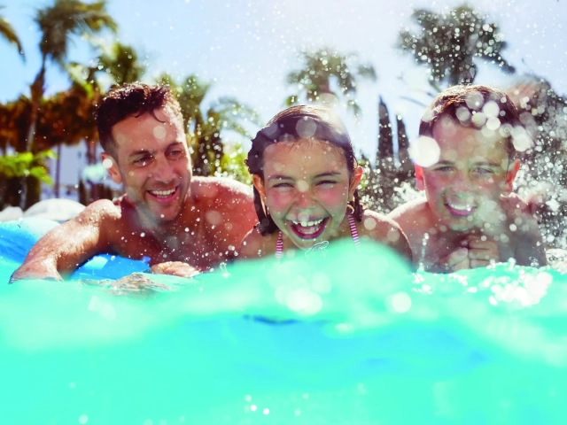 A man and two children laughing and splashing at the edge of a swimming pool, with palm trees in the background.