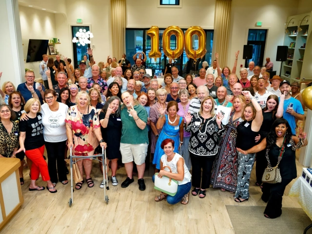 Large group of smiling, cheering people posing together indoors beneath gold balloons spelling out the number 100.