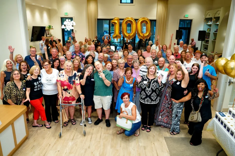 Large group of smiling, cheering people posing together indoors beneath gold balloons spelling out the number 100.