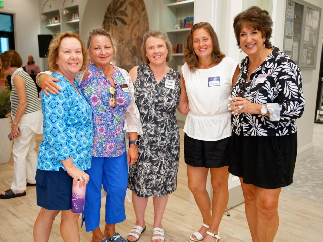 Five smiling women in colorful summer outfits standing arm-in-arm indoors, four wearing name tag stickers.