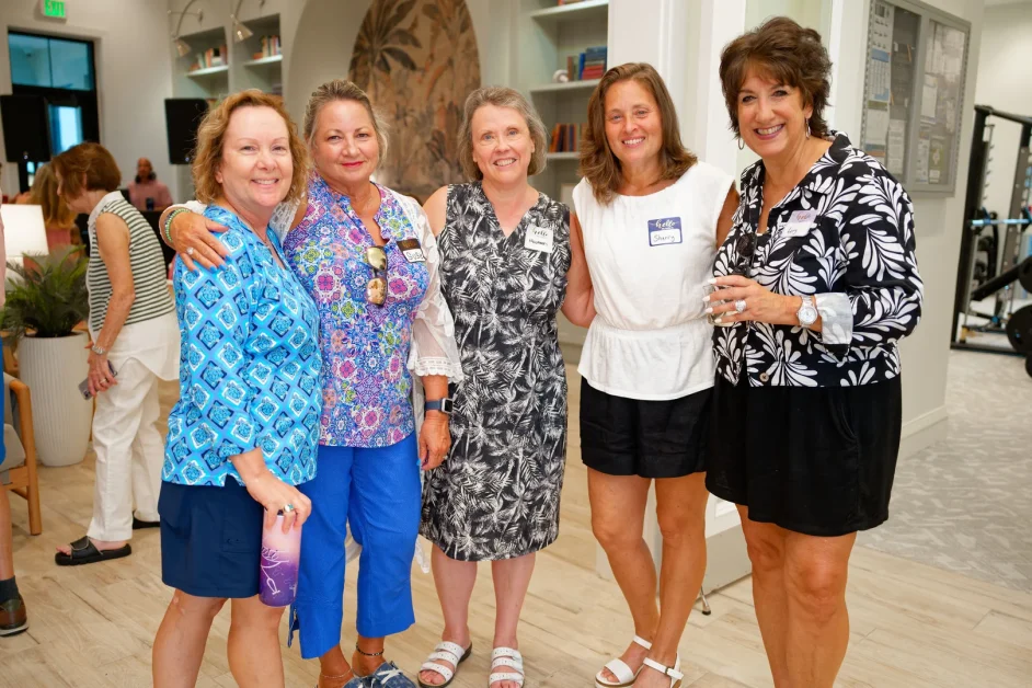 Five smiling women in colorful summer outfits standing arm-in-arm indoors, four wearing name tag stickers.