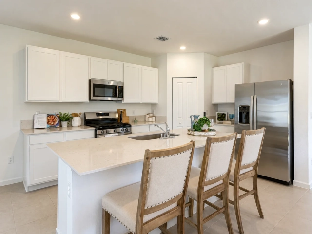 White kitchen with a light quartz island, three upholstered bar stools, and stainless steel refrigerator and range.